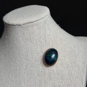 May include: An oval-shaped brooch with a gold-colored frame. The brooch features a polished, dark blue and green stone. The jewelry is displayed on a neutral-colored mannequin bust, set against a dark background.