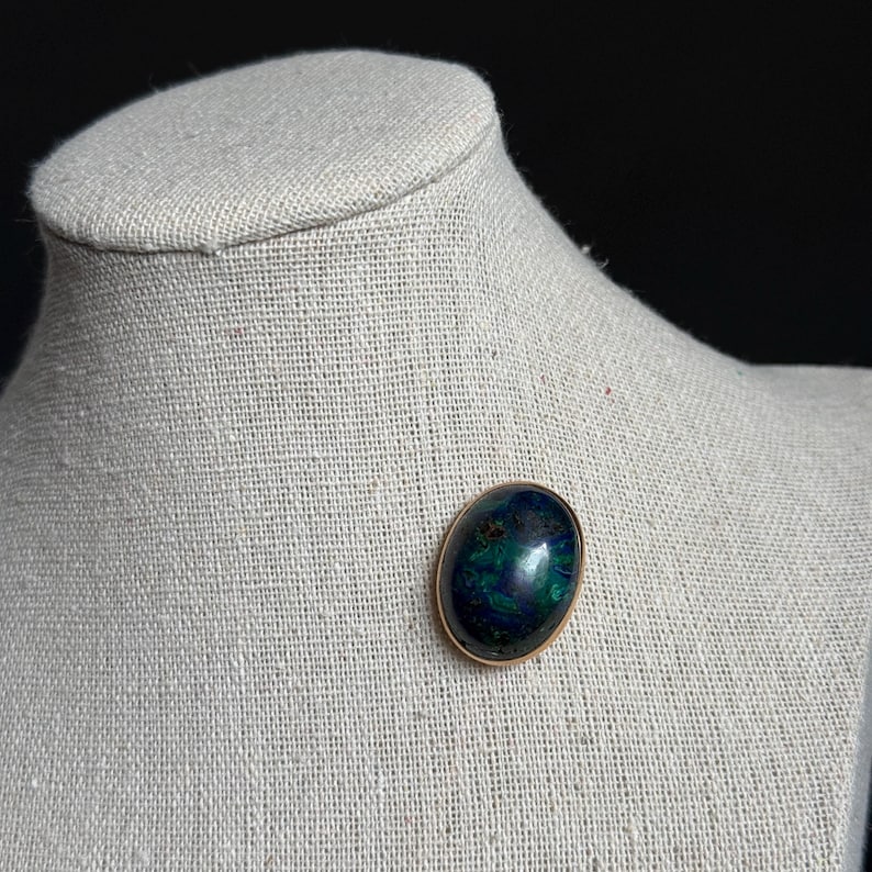 May include: An oval-shaped brooch with a gold-colored frame. The brooch features a polished, dark blue and green stone. The jewelry is displayed on a neutral-colored mannequin bust, set against a dark background.