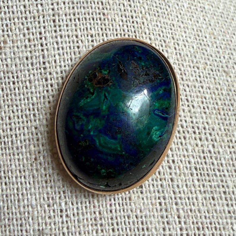 May include: Oval-shaped pendant with a gold-colored bezel. The stone features a swirling pattern of deep blue and green hues. The polished surface reflects light, highlighting the natural variations in the stone's color and texture. The pendant is set against a neutral background.