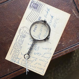 Op de afbeelding: Antieke zilverkleurige vergrootglas met een decoratief handvat, rustend op een vintage ansichtkaart met handgeschreven tekst. De ansichtkaart ligt op een bruin leren oppervlak. Het vergrootglas heeft een kleine lus aan het uiteinde van het handvat.