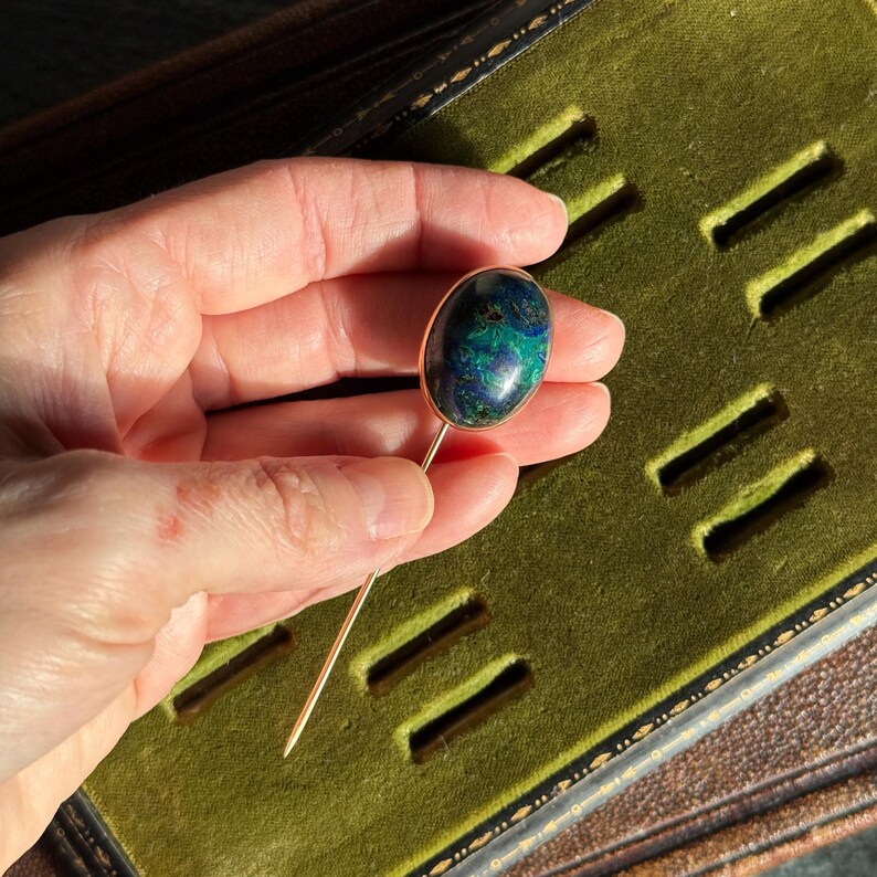 May include: A gold-toned stick pin with an oval-shaped, blue and green stone. The pin is held against a green velvet surface, showcasing the jewelry's design and color contrast. The stone appears to be approximately 0.75 inches in length.