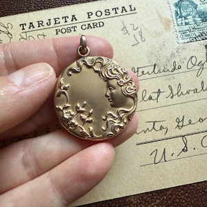 May include: A gold-colored round pendant with a woman's profile and floral details. The pendant is held in a hand, with a vintage postcard in the background. The postcard has the text "TARJETA POSTAL POST CARD".