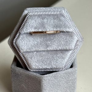 Op de afbeelding: Een dunne ring van roségoud met een delicaat gegraveerd sterrenpatroon op het oppervlak, stevig geplaatst in de met fluweel beklede gleuf van een zeshoekig lichtgrijs ringdoosje met het bijpassende deksel erbovenop.