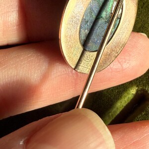 May include: Close-up of a gold-toned brooch with an oval-shaped, iridescent stone. The brooch has a pin backing and the text "OVER" is visible. The brooch is held in a hand.