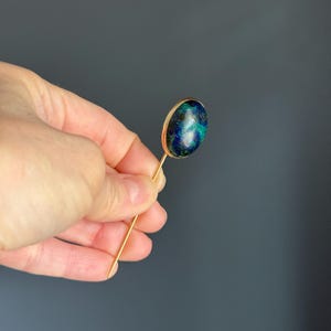 May include: A gold-toned stick pin featuring an oval-shaped stone with a mix of blue and green hues. The stone is set in a gold bezel, and the pin is held in a hand against a gray background. The pin is a jewelry item.