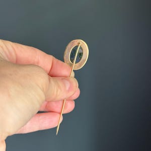 May include: A gold-toned hat pin with an oval-shaped head. The head features a dark blue and green stone set within a gold-colored frame. The pin is held against a dark gray background.