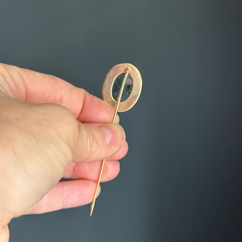 May include: A gold-toned hat pin with an oval-shaped head. The head features a dark blue and green stone set within a gold-colored frame. The pin is held against a dark gray background.