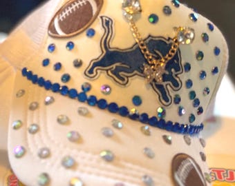Może przedstawiać: Biała czapka typu trucker z logo Detroit Lions, akcentami piłkarskimi oraz niebieskimi i przezroczystymi kryształkami. Czapka ma siatkową tylną część i zakrzywiony daszek z niebieskim obszyciem z koralików.