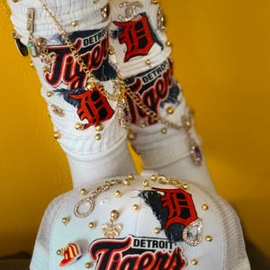 May include: White baseball cap with a red and black Detroit Tigers logo, gold chain, and rhinestones. The cap has a mesh back and is decorated with various charms.