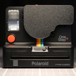 Peut inclure: Appareil photo instantané Polaroid OneStep+ noir avec un bouton rouge et un ruban arc-en-ciel. L'appareil photo est doté d'un flash intégré et d'un cache d'objectif. Le texte "OneStep+" et "Polaroid" est visible sur l'appareil photo.
