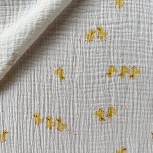 May include: White muslin fabric featuring a repeating pattern of yellow ducklings. The fabric has a crinkled texture and a folded edge. The ducklings are in various poses, adding a playful touch to the textile.