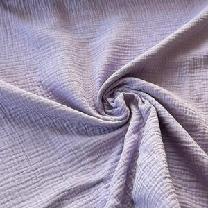 Könnte beinhalten: Nahaufnahme eines lavendelfarbenen Stoffes mit strukturierter, geknitterter Optik. Der Stoff ist gerafft und gewirbelt, wodurch Falten und Schatten entstehen, die seine weiche Textur hervorheben. Die Farbe ist ein gedämpfter Pastellton von Lila.