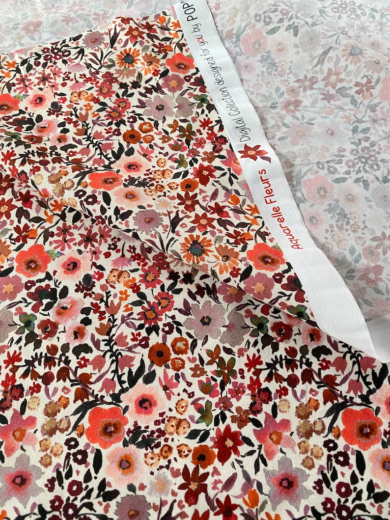 May include: Floral fabric featuring a cream backdrop and a pattern of red, orange, pink, and burgundy flowers. The fabric is folded, revealing a second fabric with a similar floral pattern. The text "Aquarelle Fleurs" and "Digital Collection designed for you by POP" is visible.