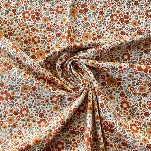 以下が含まれることがあります： オレンジ、ブラウン、イエローの色合いの小さな花のプリントが施された白い生地。生地は折り畳まれ、ドレープされており、パターンが示されています。