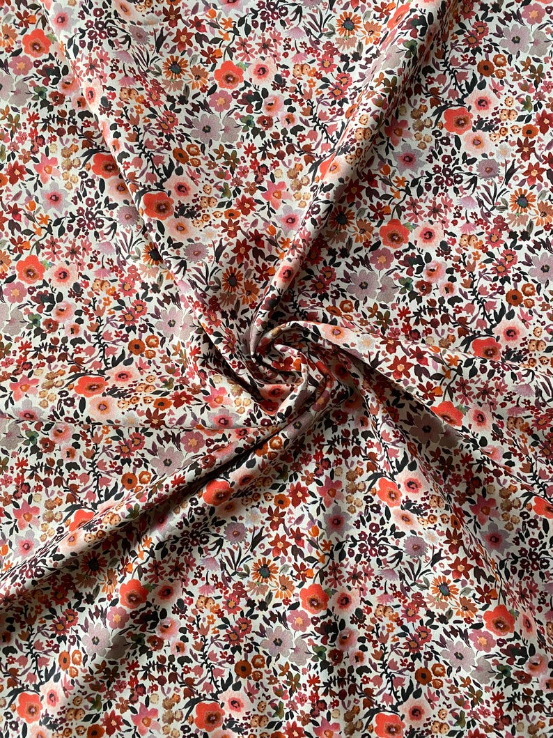 May include: A close-up of a floral fabric featuring a dense pattern of small flowers in shades of red, orange, pink, and brown. The fabric is white with a slight sheen, and the flowers are arranged closely together.