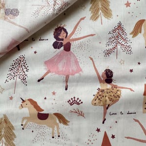 May include: White fabric with a repeating pattern of ballerinas, unicorns, crowns, and Christmas trees in shades of pink, brown, and gold. The text "Love to dance" is visible. The fabric is suitable for children's crafts.