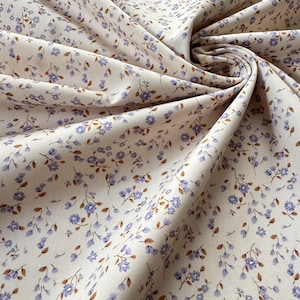 May include: A piece of off-white fabric featuring a repeating pattern of small purple flowers with brown stems and leaves. The material is draped in soft folds, creating a textured surface that highlights the delicate floral print across the textile.