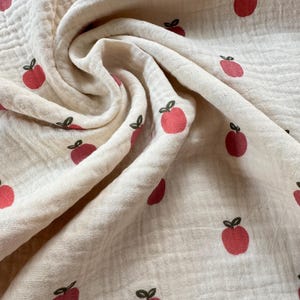 May include: Cream-colored fabric with a repeating pattern of red apples with green stems. The fabric is soft and textured, with visible folds and wrinkles. The apples are evenly spaced across the surface.