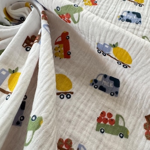 May include: White muslin fabric featuring a repeating pattern of cartoon trucks in various colours. The trucks carry items like fruit and milk. The fabric is soft and textured, with the trucks in shades of red, green, blue, and brown.
