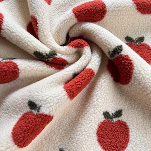 Könnte beinhalten: Nahaufnahme eines cremefarbenen Fleece-Stoffs mit einem sich wiederholenden Muster aus roten Äpfeln. Jeder Apfel hat ein grünes Blatt und einen Stiel. Der Stoff ist weich und strukturiert, mit einem gemütlichen, plüschigen Aussehen.