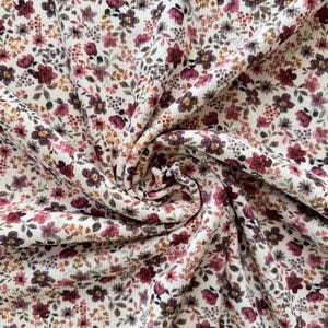 May include: Close-up of a cream-coloured fabric with a floral pattern. The fabric features a repeating design of small, burgundy, pink, and brown flowers and leaves. The fabric is textured and appears to be a knit or ribbed material.