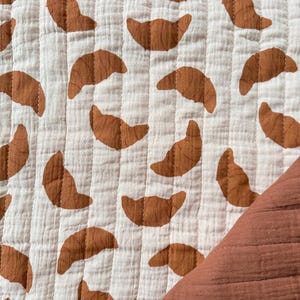 May include: A white and brown quilted fabric featuring a repeating pattern of brown croissants. The fabric has a textured appearance and is stitched with vertical lines. A corner of a solid brown fabric is visible.