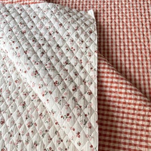 May include: A reversible quilted fabric featuring a white side with a small red and pink floral print and a red and white gingham check side. Both sides have a diamond-shaped stitched pattern throughout the textile surface.