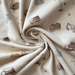 Könnte beinhalten: Cremefarbener Rippstoff mit einem sich wiederholenden Muster von Baufahrzeugen in Braun-, Grau- und Gelbtönen. Das Design umfasst Lastwagen, Betonmischer, Kräne und Bagger. Der Stoff ist eng gewebt und hat eine weiche Textur.