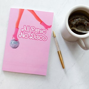 May include: A pink notebook with the text "All Panic No Disco" and a graphic of a doll's legs and a disco ball. A gold pen with a glitter barrel and a white mug with tea are also in the image.