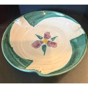 May include: A round ceramic bowl with a white interior and a hand-painted purple flower with a yellow center. The rim features a wide, textured teal green border that sweeps across the edges, creating a contrast against the off-white base.