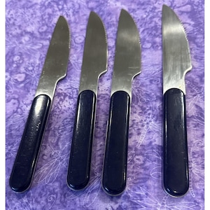 Puede incluir: Cuatro cuchillos de acero inoxidable con bordes dentados y mangos azul oscuro están colocados sobre una superficie morada estampada. Los cuchillos tienen un diseño clásico, adecuados para diversas tareas de cocina.