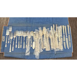 Pode incluir: Uma grande coleção de talheres em tom prateado, incluindo garfos, colheres e facas, dispostos num rolo de tecido azul. Muitos dos utensílios estão embalados individualmente em mangas de plástico transparente com cabos decorados.