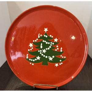 May include: A round, glossy red ceramic plate featuring a centered graphic of a dark green Christmas tree. The tree is decorated with white star shapes, small white circular ornaments, and vertical white candle illustrations throughout.