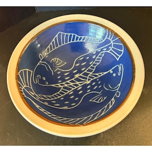 May include: A round ceramic bowl featuring a deep blue interior with a white line-art illustration of two fish swimming in a circular pattern. The bowl has a wide, off-white rim and a brown-toned edge separating the rim from the blue center.