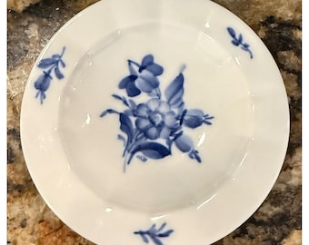 May include: A white porcelain plate featuring a hand-painted blue floral motif in the center. The design includes a central bouquet of flowers and leaves, with two smaller sprigs positioned on the rim. The plate has a smooth, glossy finish.
