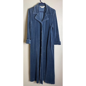 May include: A long, blue velvet robe with a collared neckline and long sleeves. The robe is buttoned down the front and has pockets. The sleeves are cuffed at the wrists. The robe is hanging on a black hanger against a neutral background.