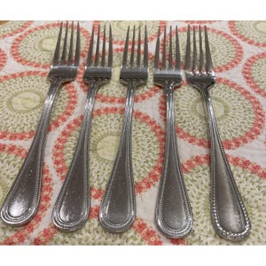 May include: Five silver forks with a decorative beaded edge and a teardrop-shaped handle are arranged on a patterned tablecloth. The forks have four tines each and are made of a shiny metal.