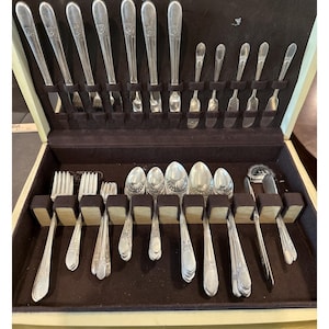 Op de afbeelding: Een complete set vintage bestek, inclusief vorken, lepels, messen en serveerbestek, netjes gerangschikt in een houten kist met een donkerbruine fluwelen voering. Het bestek heeft een decoratief bloemmotief.