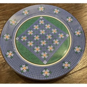 May include: A decorative plate with a blue and green geometric design. The plate features a central green circle with a diamond pattern, surrounded by a blue border with a grid pattern and square accents. The plate has a gold rim.