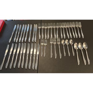 May include: A large collection of silver-toned stainless steel flatware arranged in rows. The set includes numerous dinner knives with serrated blades, dinner forks, salad forks, and oval-shaped spoons, all featuring a simple, uniform handle design.