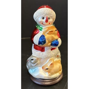 May include: A snowman figurine with a red hat and coat, holding a golden rabbit. The snowman has a green scarf and blue mittens. Two rabbits are at the base of the snowman. The figurine is on a brown base.