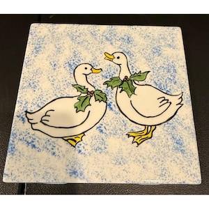 May include: A square ceramic tile featuring two white ducks with yellow beaks and feet. Each duck wears a green holly leaf collar with small red berries. The background is a white surface with a light blue speckled or sponge-painted texture.