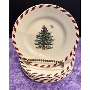 May include: A stack of white ceramic plates featuring a central illustration of a decorated Christmas tree with gifts underneath and a Santa figure on top. The rim of each plate is decorated with a repeating red and white diagonal candy cane stripe.