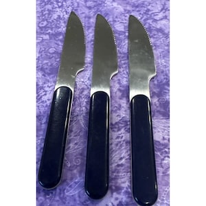 Puede incluir: Tres cuchillos de carne de acero inoxidable con hojas dentadas y mangos azul marino. Los cuchillos están dispuestos verticalmente sobre un fondo morado estampado. Las hojas son brillantes y reflejan la luz.