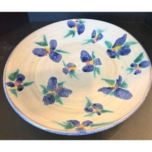 Pode incluir: Um prato redondo de cerâmica com uma base de cor creme apresentando um padrão pintado à mão de flores de íris azuis com folhas verdes e centros amarelos. A borda do prato tem uma textura azul salpicada e o design está distribuído pela superfície.