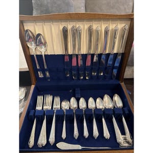 May include: A silver-toned cutlery set presented in a wooden storage chest with a blue velvet interior. The collection comprises various spoons, forks, and knives, each displaying a decorative floral pattern on the handle. A single butter knife is positioned horizontally at the base of the case.