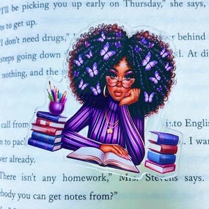 May include: A sticker featuring a Black woman with purple butterfly hair, wearing glasses and a purple striped shirt, reading a book. The sticker has a stack of books and a pencil holder. The background is a book page.