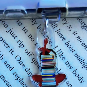 May include: A clear acrylic keychain featuring a stack of colorful books with a red heart and a red hand holding a pen design. The keychain hangs from a silver-colored chain. The background is a page of text.