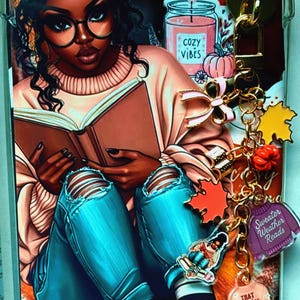 May include: Clear phone case featuring an illustration of a person reading a book, wearing glasses, a pink sweater, and ripped blue jeans. The case has a gold charm with fall-themed charms and the text "Sweater Weather Reads" and "That Sweater Weather Feeling."