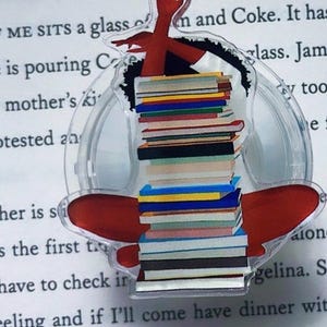 May include: A clear, circular bookmark featuring a stack of colorful books with a red figure on top. The bookmark is placed on a book page with printed text.